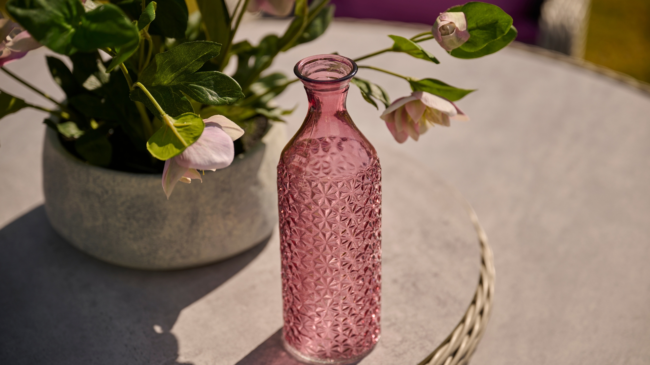 pink glass vase on a table top with flowers in the background 