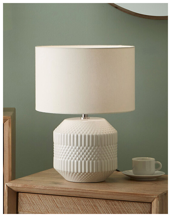 White Embossed Geo Ceramic Table Lamp set on a wooden chest of drawers.