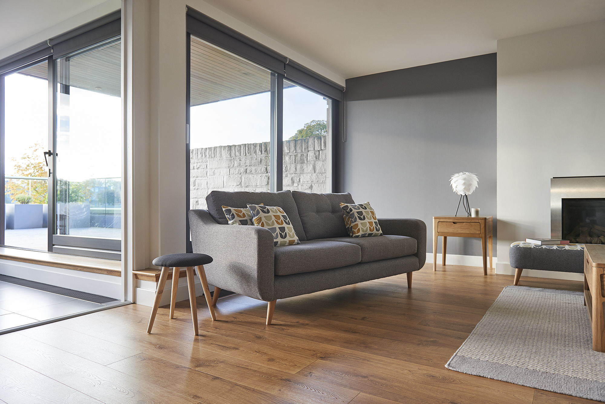 Mid century modern sofa in grey fabric with bright scatter cushions shown against a dark wall, wooden floor and a big window